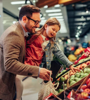 Little girl is buying groceries in the supermarket with her father. He carrying her and picking together fresh fruits